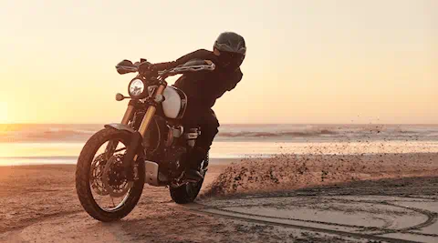 An image of a stunt rider riding a Scrambler 1200 in the sand on a sunset beach.